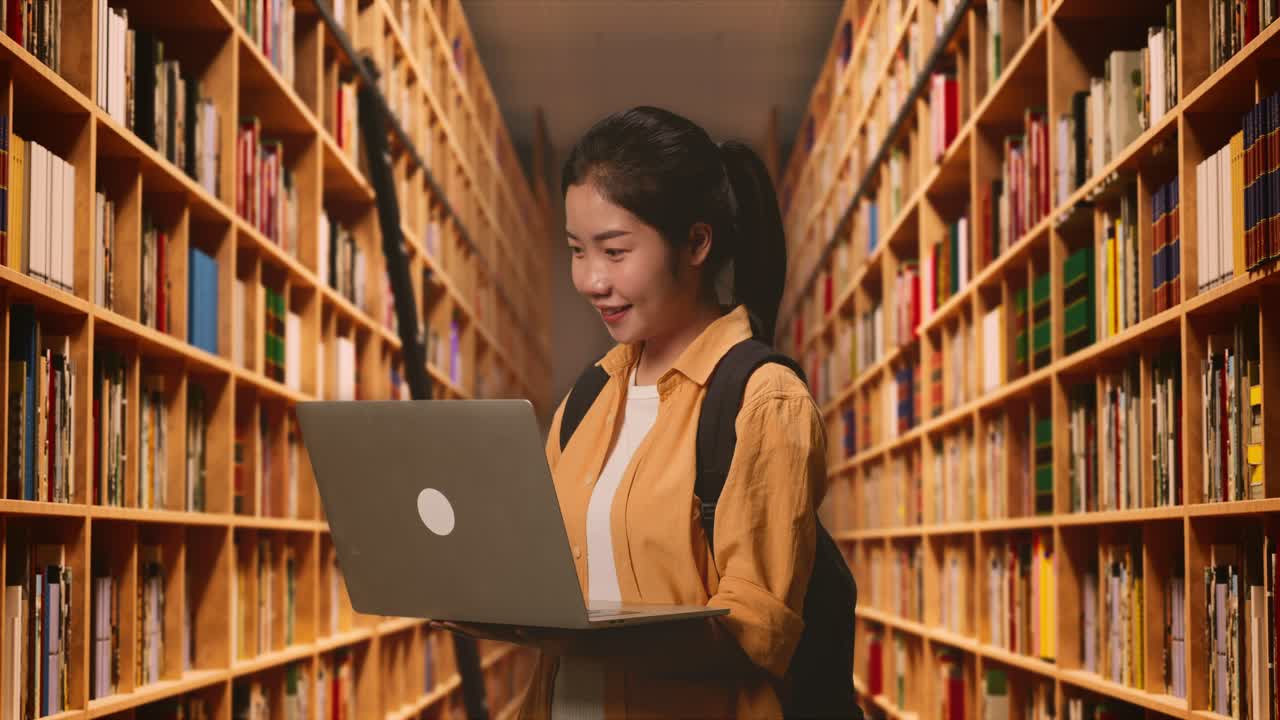 Side View Of Asian Woman Student With A Backpack Smiling And Typing On A Laptop While Standing In The Library
