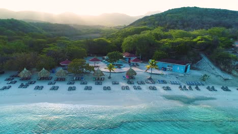 Grote-Knip-beach-at-sunrise,-golden-glow-spreads-across-ocean-water-and-mountains-behind,-Curacao