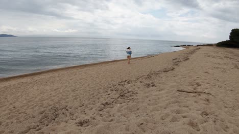 A-young-girl-walking-on-a-beach-in-Greece,-on-a-partly-cloudy-day,-untying-her-brown-hair