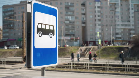 vista cercana de una señal de parada de autobús en un entorno urbano con peatones caminando en el fondo, mientras que algunas personas sólo están de pie