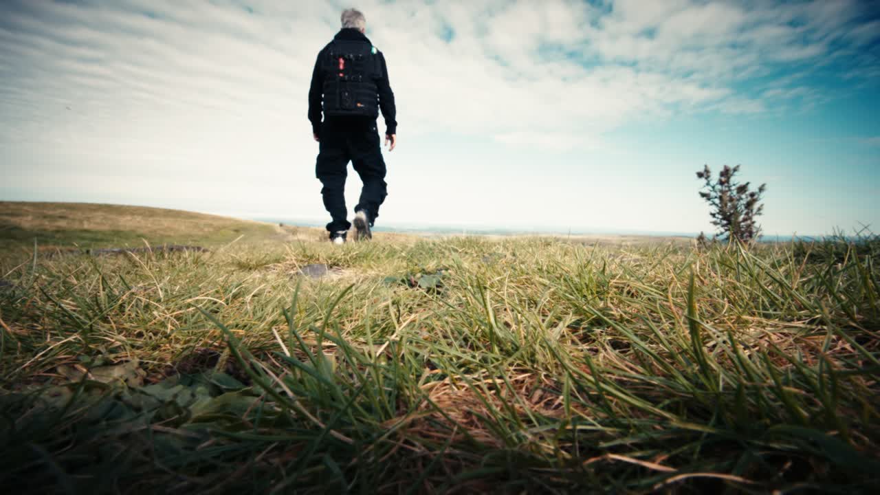 Låg vinkelvy av en fotograf med ryggsäck stående på en gräsklädd kulle mot en molnig himmel. Markperspektivet framhäver silhuetten och den naturliga miljön
