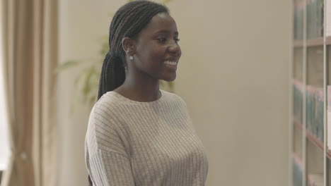 Una-Linda-Mujer-Negra-Toma-Un-Libro-En-La-Biblioteca-Y-Sonríe-Directamente-A-La-Cámara