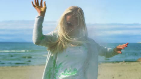 beautiful young young girl with blonde hair wearing cool sunglasses is hit by a colorful holi powder, throws some in return and laughs. clear blue sky behind her and deep blue sea behind her.