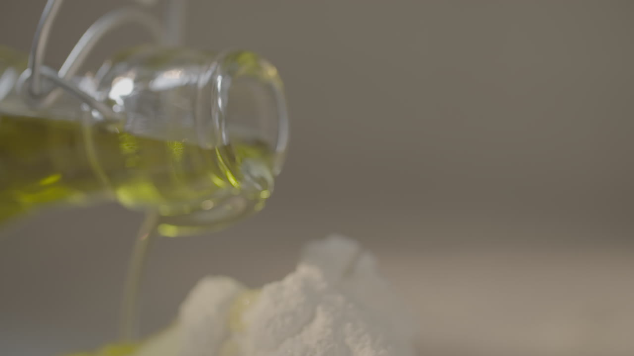 Close up slowmotion shot of a glass oil bottle pouring olive oil on top of some baking flour dripping all over LOG