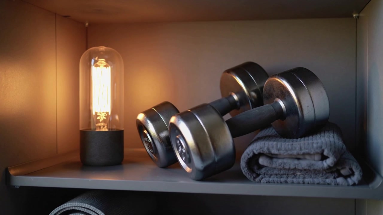Moving camera shifting over home gym shelf, showing lamp glow and stacked dumbbells on towel
