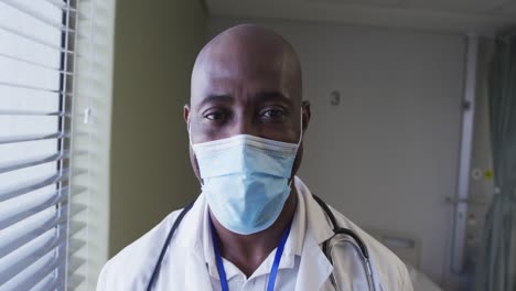 Portrait-of-african-american-male-doctor-wearing-face-mask-standing-in-hospital-room