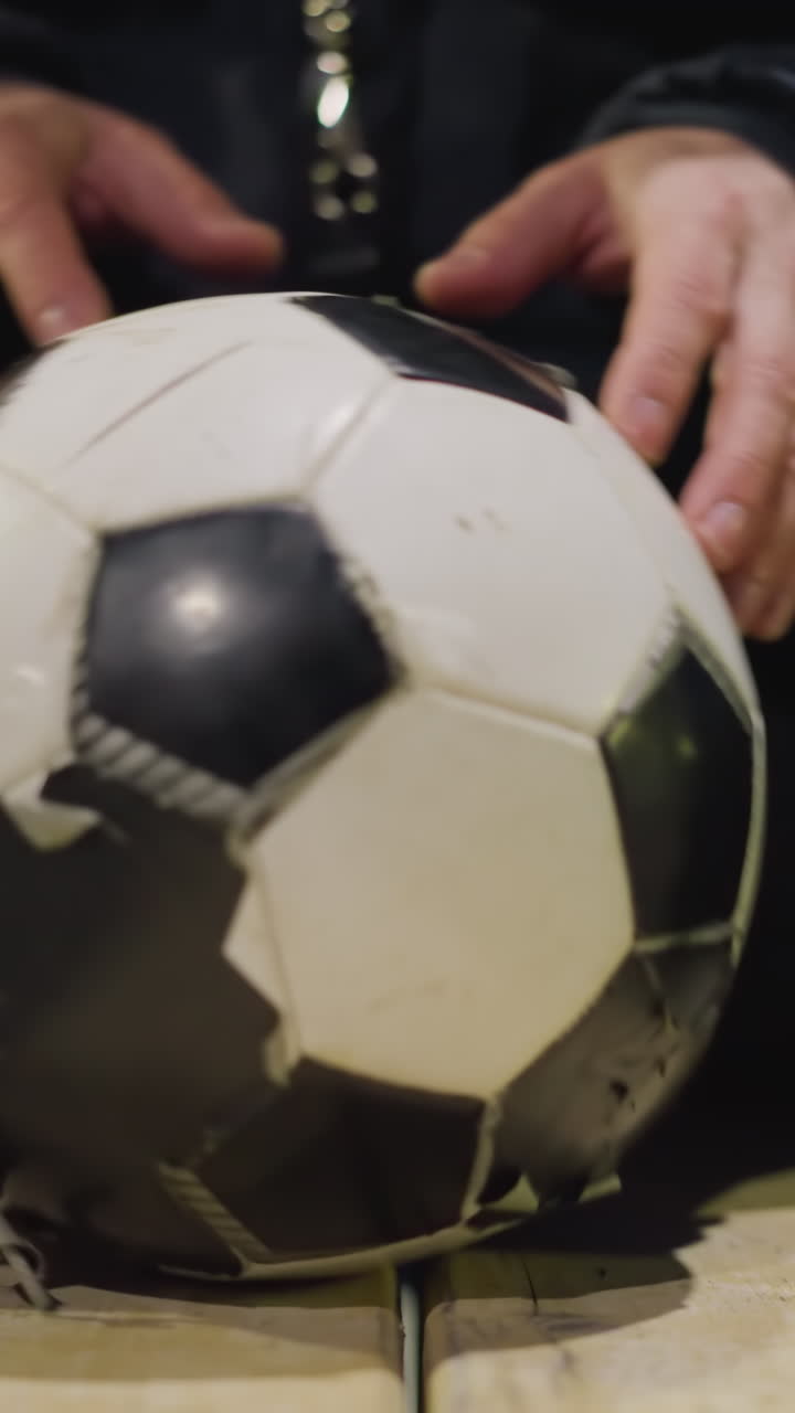 Close-up of a person sitting on a bench with a soccer ball between their legs, gently rotating the ball with his hands with blur dimly lit background