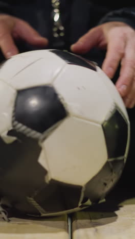 close-up of a person sitting on a bench with a soccer ball between their legs, gently rotating the ball with his hands with blur dimly lit background