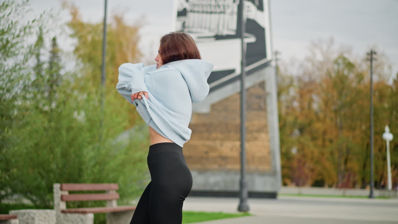 Close-up view of lady removing her blue sweater in park with background featuring buildings, poles, and trees. Ideal for wellness, outdoor, and casual lifestyle moments in serene environment