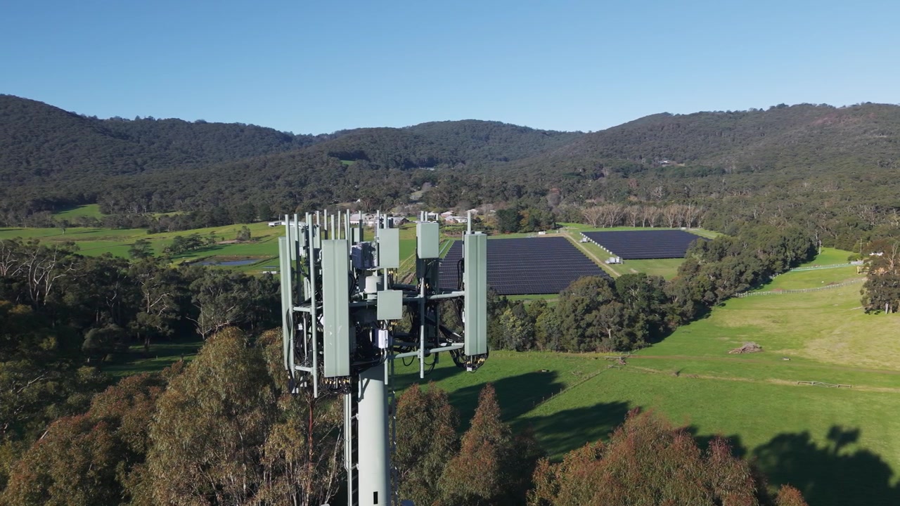 Video con drone che sorvola un'alta torre cellulare che domina campi lussureggianti e un grande parco solare a The Basin, Melbourne. La luce diurna nitida, l'ampio paesaggio e i cieli sereni offrono una tranquilla scena rurale.