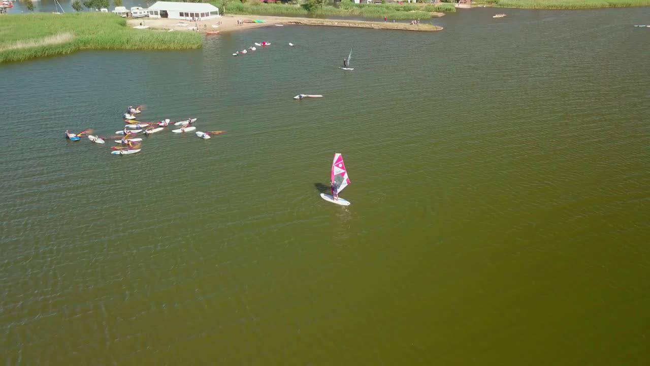 rodeando la toma aérea, la cámara se desplaza hacia arriba desde el grupo de windsurfistas ociosos, revelando el cielo despejado y soleado
