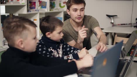 Attarctive-male-programmer-teacher-with-several-tattooes-on-his-arms-explaining-something-to-his-two-little-pupils-boys-on-a-programming-class.-Alternative-education-for-children.