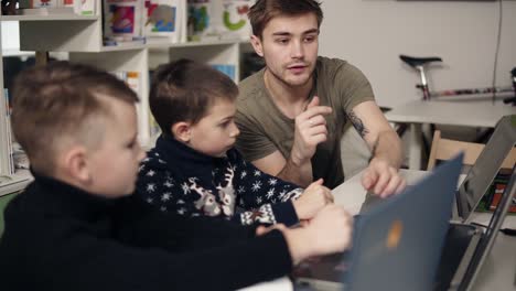 attarctive male programmer teacher with several tattooes on his arms explaining something to his two little pupils boys on a programming class. alternative education for children.