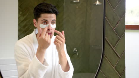 Focused-biracial-man-applying-under-eye-masks-looking-in-bathroom-mirror,-copy-space,-slow-motion