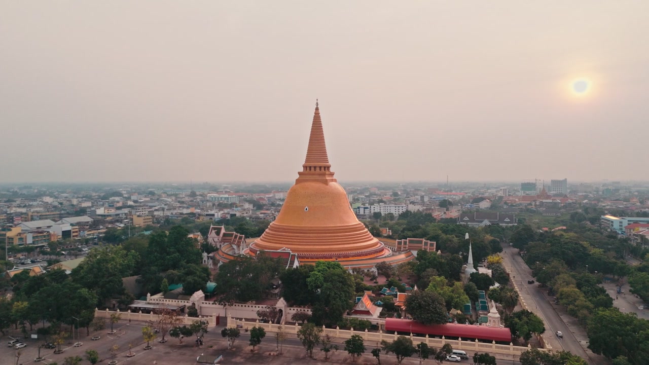 Una prospettiva aerea da drone cattura un'enorme stupa dorata che si erge sopra il centro urbano, mentre un maestoso monumento buddista domina lo skyline della città in Thailandia sotto un cielo grigio e velato con il sole mattutino splendente.