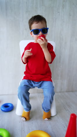 caucasian toddler wearing sun glasses sits on the chair at home. cute baby boy eating an apple. vertical video.