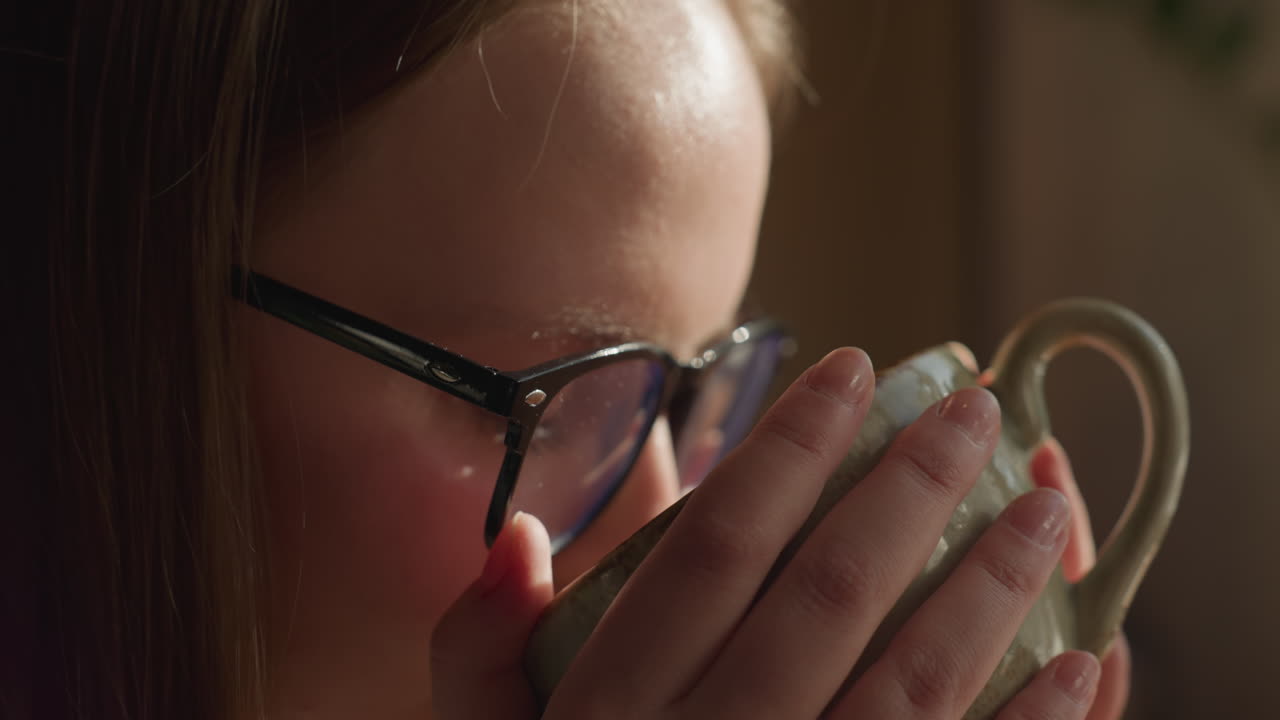 Close up of fair skin lady with glasses gently holding warm tea cup near window as soft sunlight highlights her hand, thoughtful ambiance with delicate textures of sweater and ceramic cup in focus