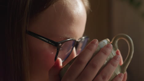 close up of fair skin lady with glasses gently holding warm tea cup near window as soft sunlight highlights her hand, thoughtful ambiance with delicate textures of sweater and ceramic cup in focus