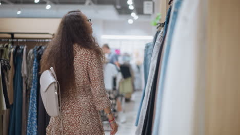 woman with long curly hair and stylish dress browsing through clothing racks in store, carrying a white backpack, other shoppers in background, creating a lively retail atmosphere with soft lighting