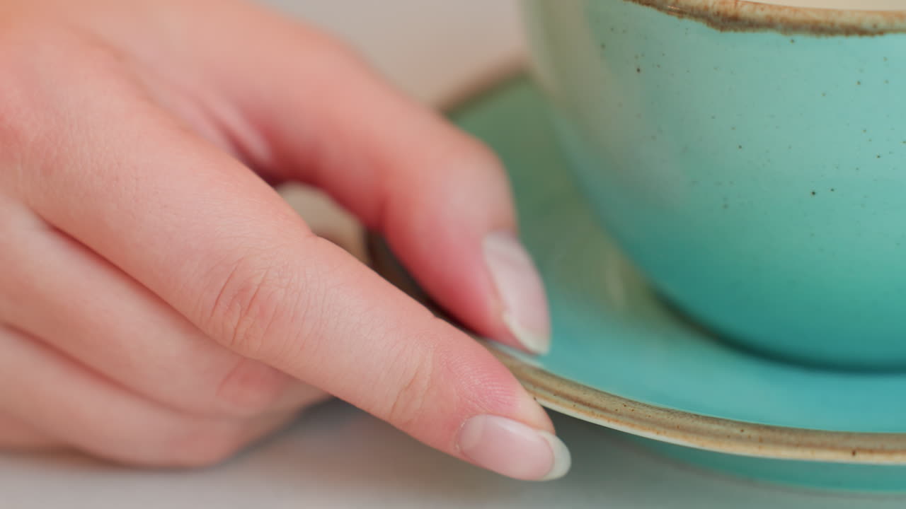 Close up of light skinned young woman adjusting her drink with tips of fingers, delicately touching rim of ceramic saucer while seated at indoor table in soft natural light creating cozy atmosphere