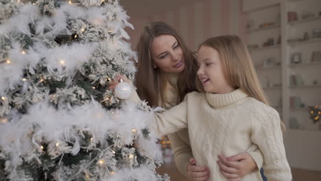 mother and daughter decorate christmas tree with white garlands and ornaments