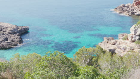 Turquoise-waters-lapping-against-a-rock-formation-adorned-with-green-plants,-creating-a-vibrant-summer-scene-in-Mallorca,-Spain