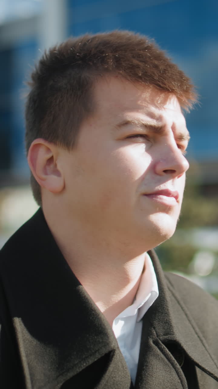 빛나는 금속 버튼을 가진 우아한 검은 코트의 클로즈업, 튼튼하게 잡힌 브리프케이스와 함께, 은 자동차, 초록색, 유리 건물을 특징으로하는 야외 도시 환경에서