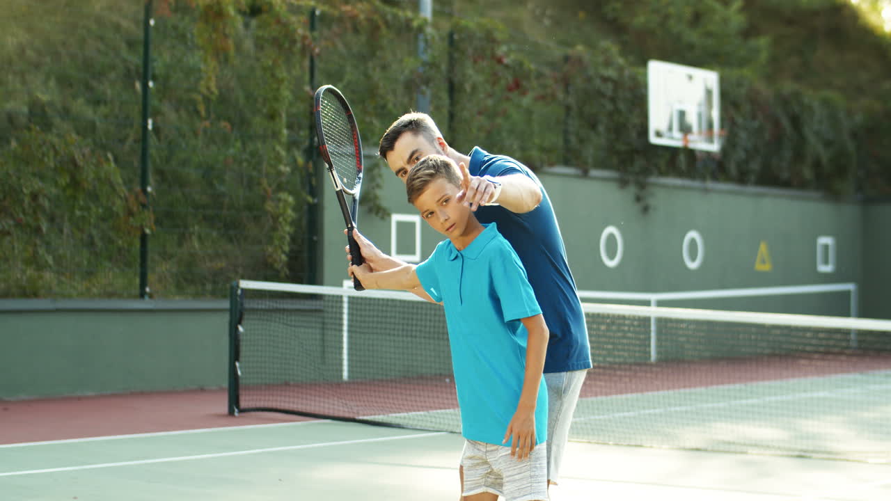 Man Teaching His Teen Son How To Play Tennis On A Summer Day 1