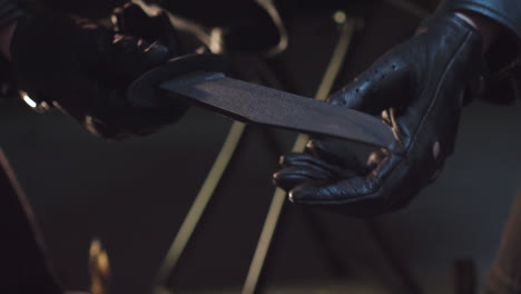 a person wearing black leather gloves holds a knife, tests its sharpness, and then places it in a holster