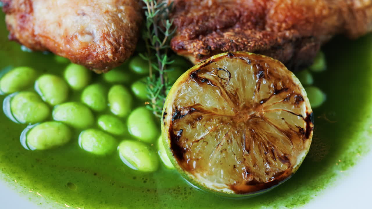 Close-up of a gourmet roasted chicken thigh served with edamame beans, a drizzle of herb-infused green sauce, and a charred lime half