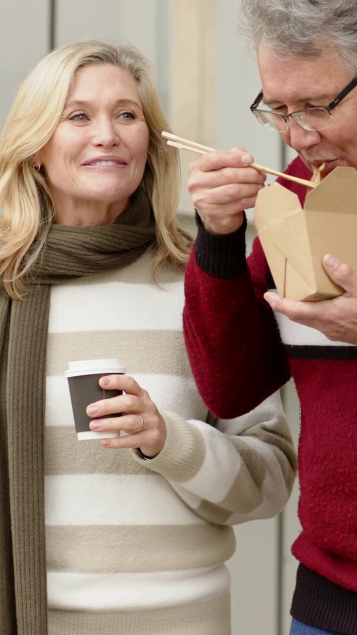 Vertical video: Holding takeout, man in red sweater eating with chopsticks near wall, woman smiling