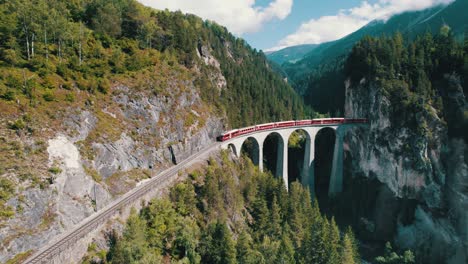 aerial view of a moving red train along the landwasser viaduct in swiss alps