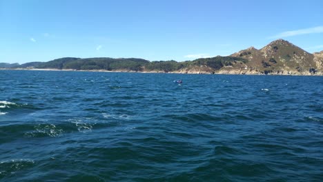 Boat-with-fishermen-sailing-fast,-the-sea-altered-near-the-Cíes-Islands-in-the-Rías-Baixas,-sunny-day-of-blue-sky-in-the-Rías-Baixas,-shot-traveling-laterally-to-the-left,-Pontevedra,-Galicia,-Spain