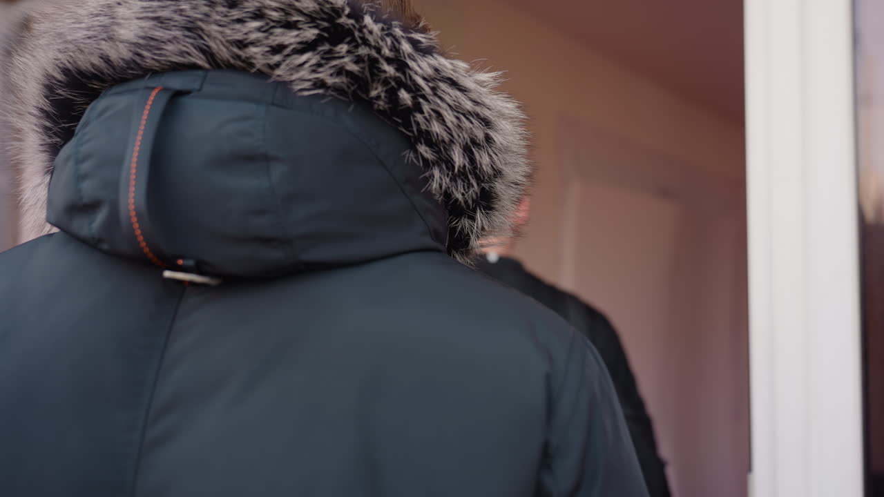 Two guys wearing jackets walks into a gym of wooden building, one with black backpack, other with fur hood coat, appearing ready to enter doorway during daylight urban scene with reflections on glass