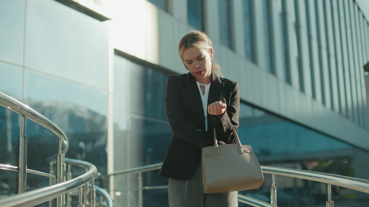 Lecturer heading home after work, attentively checking handbag to retrieve car keys, standing near modern glass building reflecting urban environment