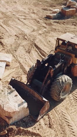 professional equipment with a bucket is turning over a stone and raising it near sandy quarry on the background of trees. Close-up. Vertical video