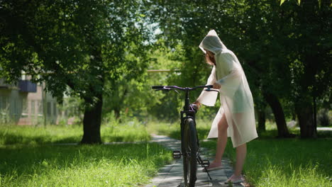 young woman in translucent raincoat stands on sunny path with her bicycle, resting her arm on handlebars and gazing thoughtfully into distance, background features lush greenery and trees