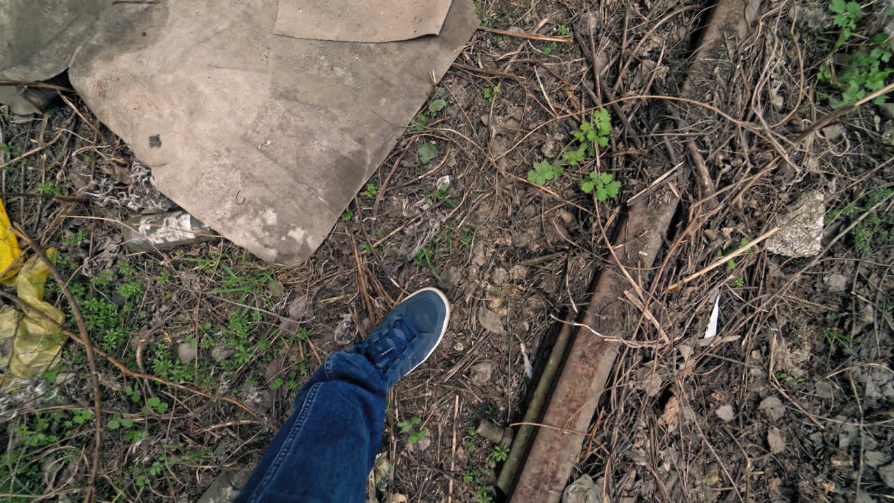 Legs of a man walking through the dirty ground with different garbage outdoors. Male's feet stepping along the dirty place with glass bottles and rubbish.
