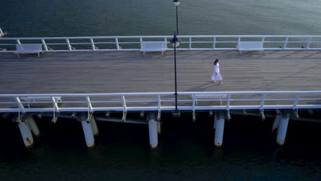 Drone-side-to-side-tracking-follows-single-woman-walking-along-pier-boardwalk-at-sunset