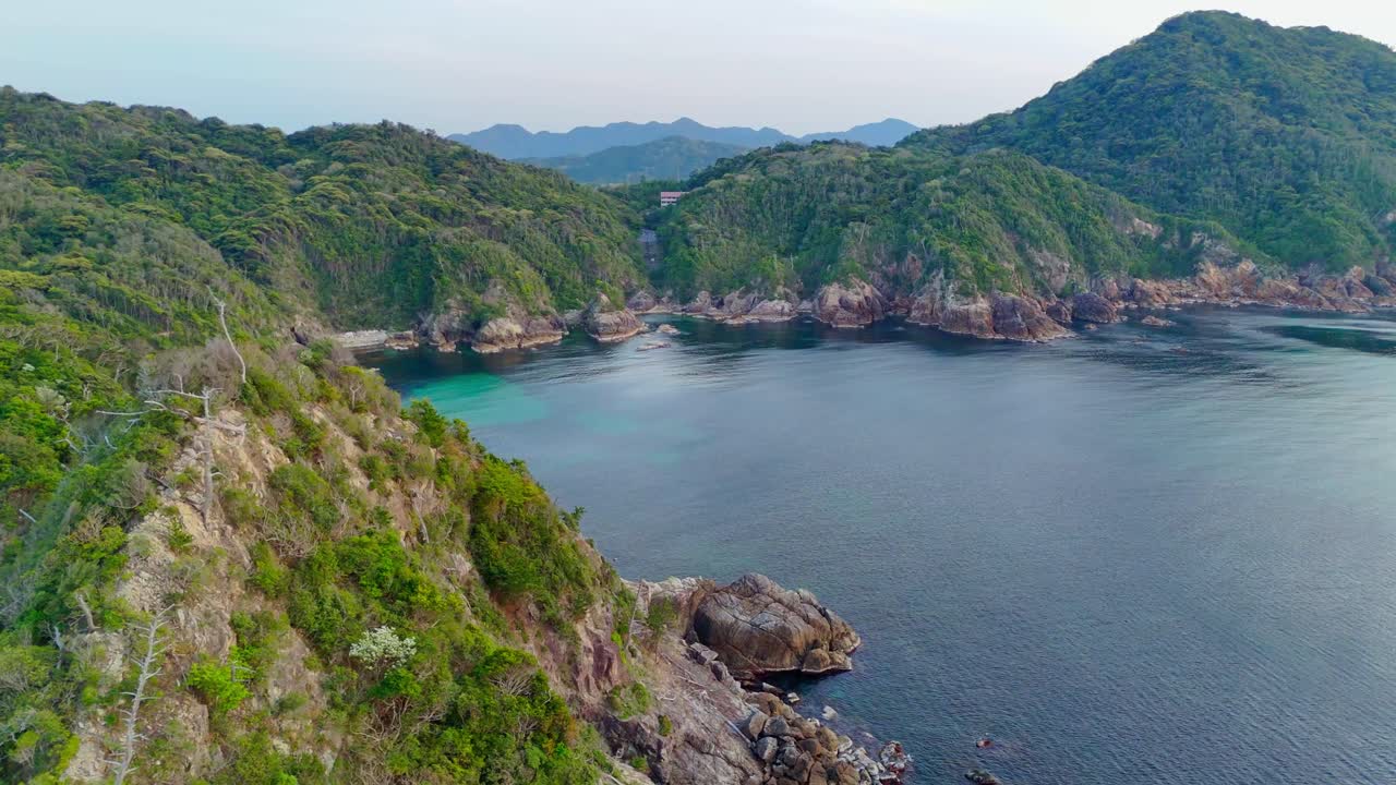 A smooth aerial flight over a tranquil bay on Japan's Uradome Coast, revealing lush green cliffs, rugged rocky shores, and patches of clear turquoise water below