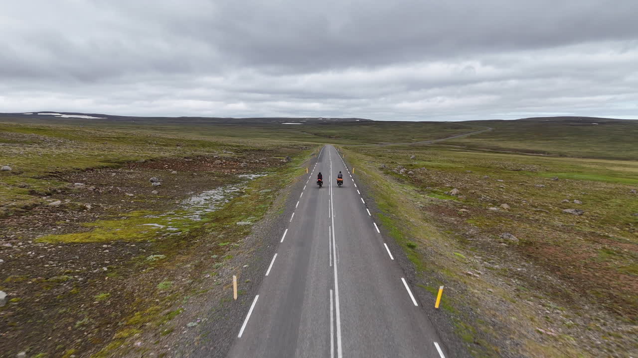 Aerial drone view of two cyclists riding along a remote coastal road in Eastern Iceland, with fjord, mountains, and open landscape under overcast skies, symbolizing adventure travel, solitude, and fre