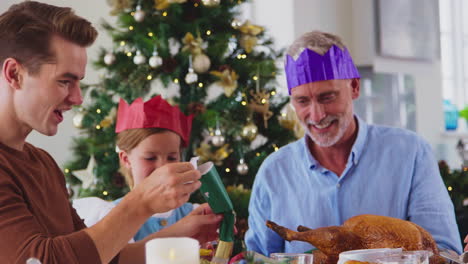 multi-generation family celebrating christmas at home wearing paper hats from crackers before meal