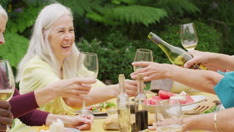 animation of diverse happy senior female and male friends eating lunch in garden, drinking wine