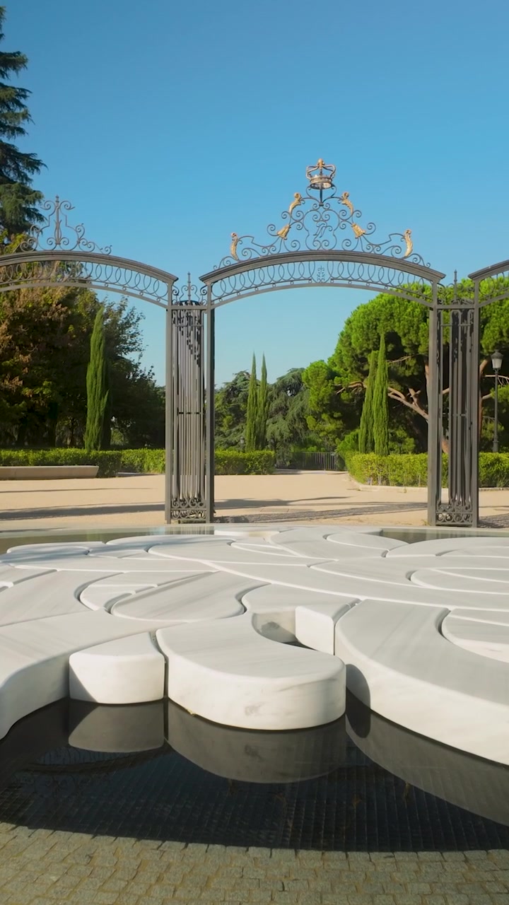 A minimalist white stone sculpture sits in a reflecting pool before historic iron gates in a sunny Spanish park. Ideal for travel, art, and wellness content.