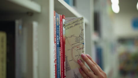 vista de la mano de la dama en vestido rosa sacando un libro de un estante en una librería, luego devolviéndolo, otros libros están ordenadamente dispuestos en el estante, con el fondo de la tienda bien iluminado y ligeramente borroso