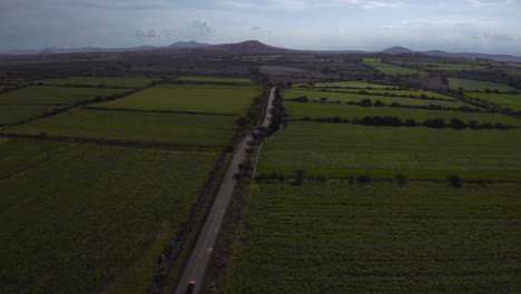 Aerial-drone-following-shot-over-a-car-passing-by-on-rural-road-through-countryside-surrounded-by-green-farmland-on-a-cloudy-day