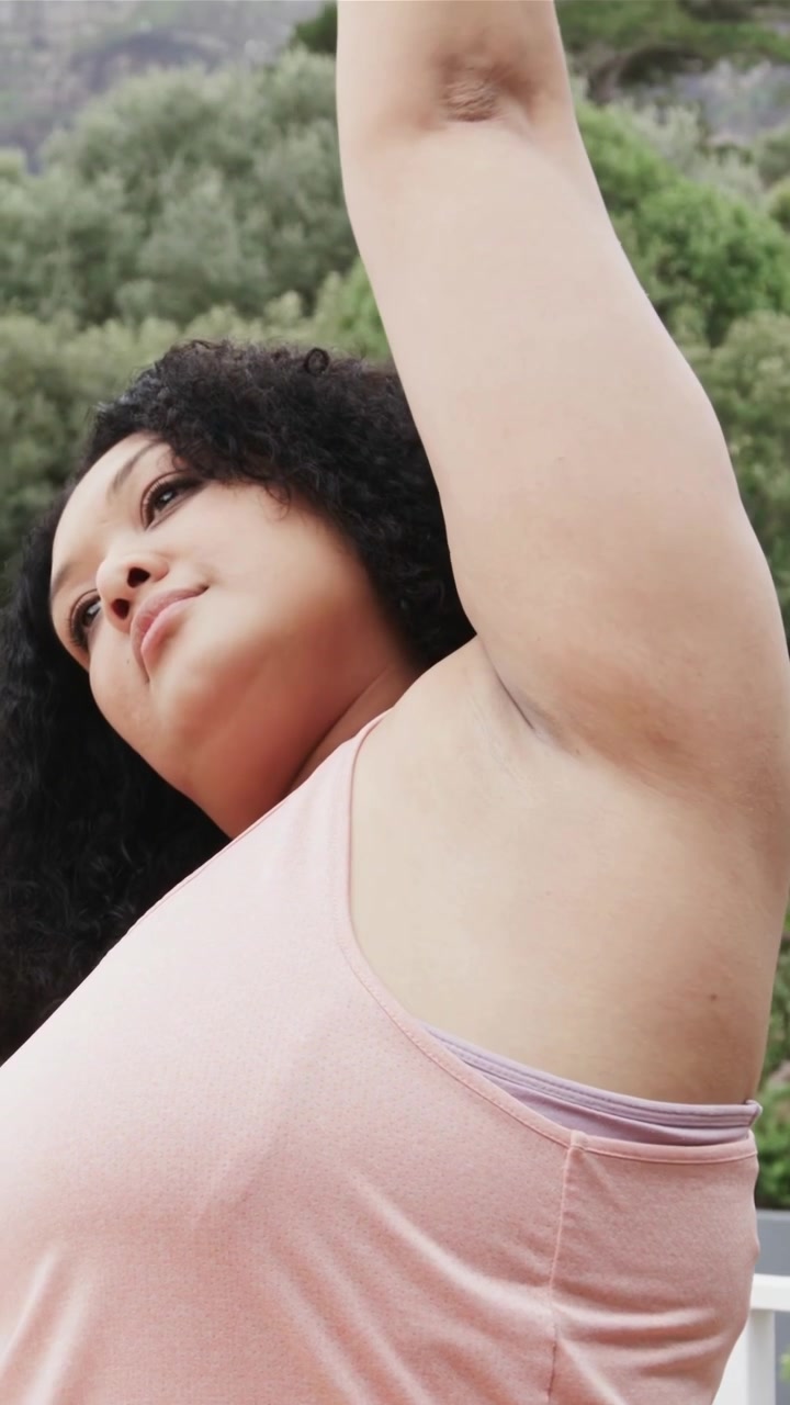 Video verticale: Donna con canotta rosa che abbassa lo sguardo e fa sollevamenti delle braccia su una piattaforma per concentrarsi, con smartwatch.