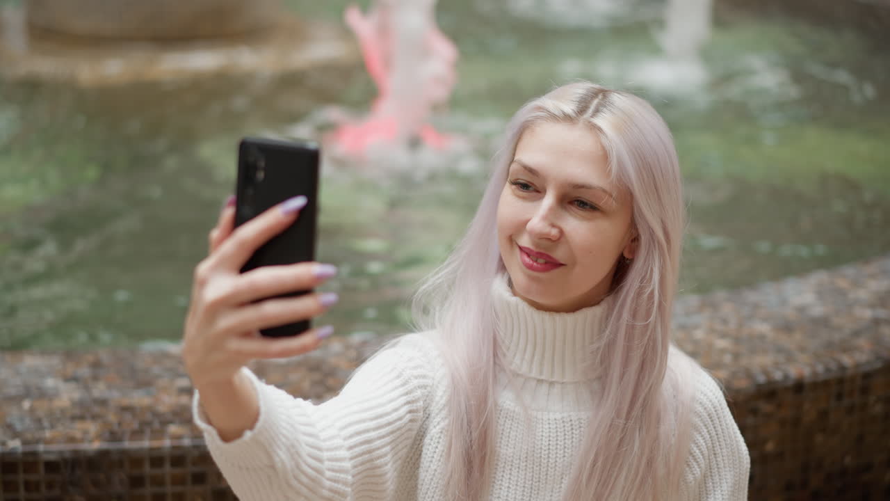 High angle view stylish trendsetter seated behind mall water fountain adjusts hair and smiles while taking selfie on mobile phone with blurred fountain backdrop and bustling mall atmosphere