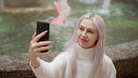 high angle view stylish trendsetter seated behind mall water fountain adjusts hair and smiles while taking selfie on mobile phone with blurred fountain backdrop and bustling mall atmosphere