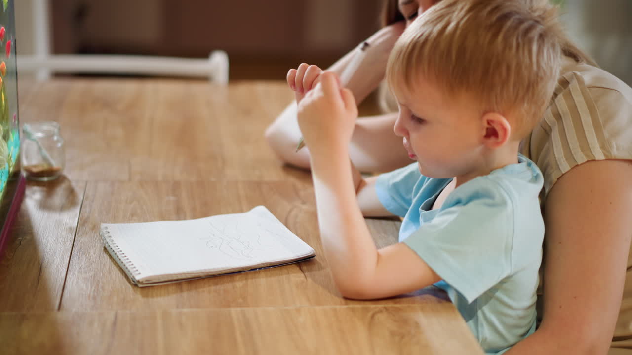 Mother supports child learning to hold pen while drawing in notebook on wooden table, showing concentration, patience, and educational guidance during early childhood development